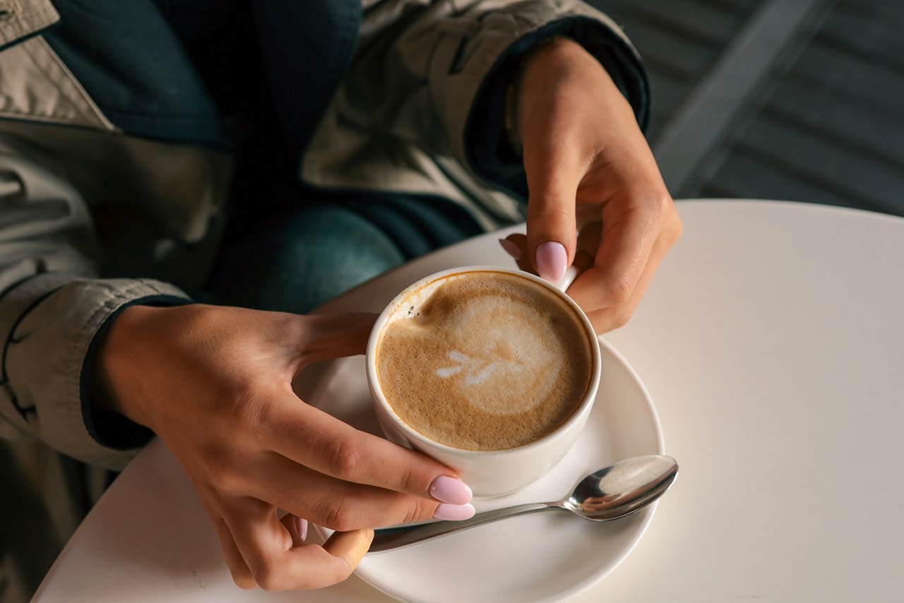 Close-up of woman's hands in trench coat gently holding a cup of cappuccino with latte art, seated at a white table